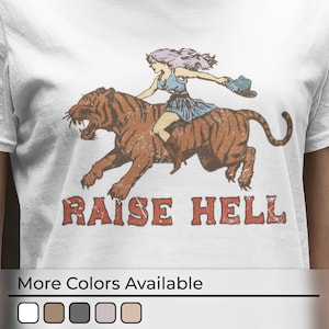 Puede incluir: Camiseta blanca con un gráfico de una mujer montando un tigre. El texto "Raise Hell" está impreso debajo de la imagen.