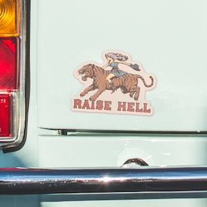 May include: A sticker with a woman riding a tiger with the text "Raise Hell" in red and white letters.