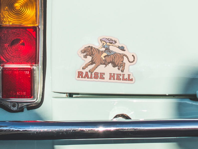 May include: A sticker with a woman riding a tiger with the text "Raise Hell" in red and white letters.