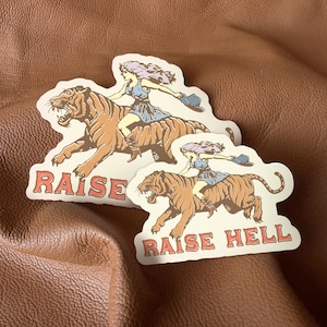 May include: Two stickers with a woman riding a tiger. The stickers are white with black outlines and feature the text "Raise Hell".