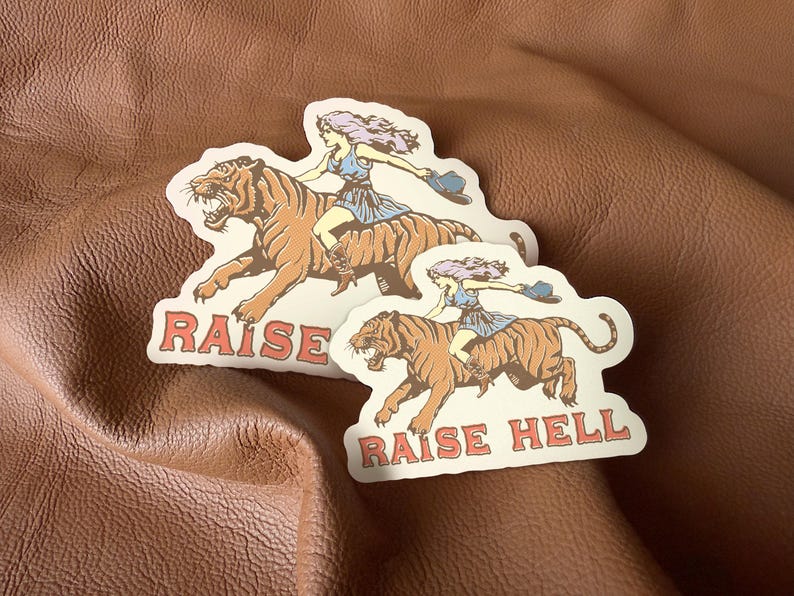 May include: Two stickers with a woman riding a tiger. The stickers are white with black outlines and feature the text "Raise Hell".
