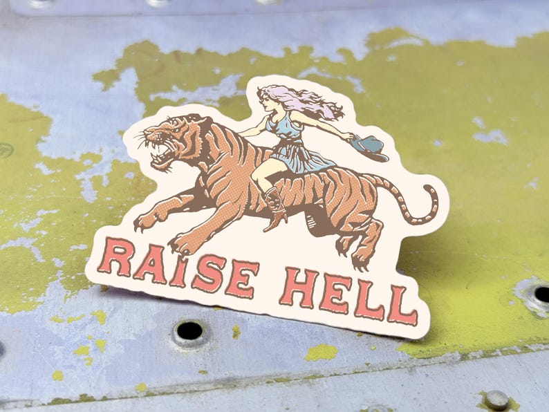 May include: A sticker featuring a woman riding a tiger with the text "RAISE HELL" in pink letters.