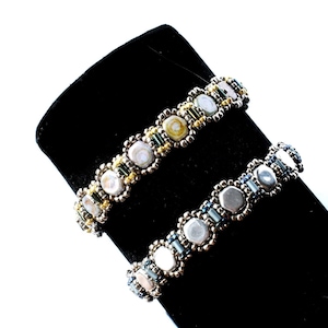 May include: Two beaded bracelets on a black display stand. The top bracelet has silver, gold, and green beads. The bottom bracelet has silver, white, and blue beads.