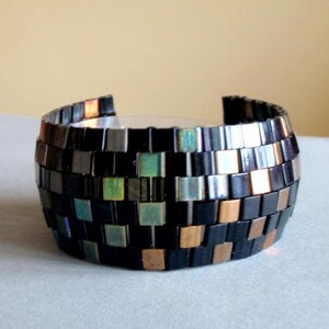 Pu&ograve; includere: Un braccialetto in mosaico nero e rame con piastrelle iridescenti. Il braccialetto &egrave; realizzato con piccole piastrelle quadrate disposte in un motivo geometrico.