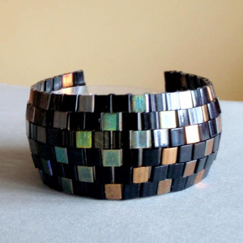 Pu&ograve; includere: Un braccialetto in mosaico nero e rame con piastrelle iridescenti. Il braccialetto &egrave; realizzato con piccole piastrelle quadrate disposte in un motivo geometrico.