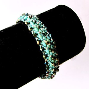 Pu&ograve; includere: Un braccialetto di perline turchesi e argento con un design intrecciato. Il braccialetto &egrave; esposto su un supporto in velluto nero.