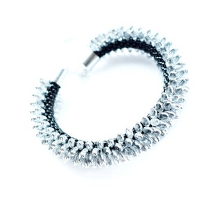 May include: A silver and black beaded bracelet with a spike design. The bracelet is made with small, silver beads that are arranged in a spike pattern. The bracelet is closed with a silver clasp.