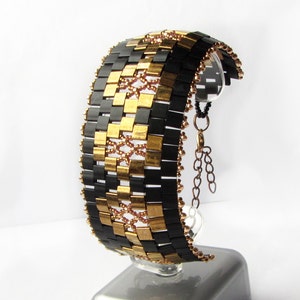 Op de afbeelding: Een zwarte en gouden kralen armband met een kettingsluiting. De armband is gemaakt van kleine, vierkante kralen die in een geometrisch patroon zijn gerangschikt.