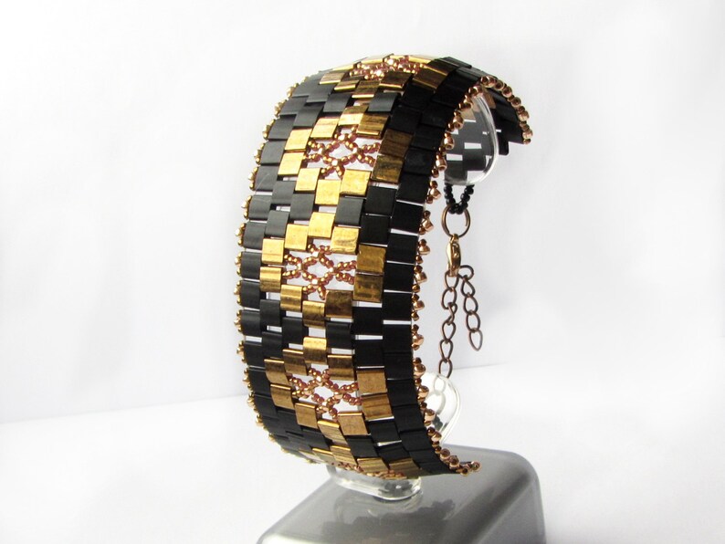 Op de afbeelding: Een zwarte en gouden kralen armband met een kettingsluiting. De armband is gemaakt van kleine, vierkante kralen die in een geometrisch patroon zijn gerangschikt.