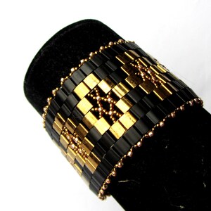 Op de afbeelding: Een zwarte en gouden kralen armband met een geometrisch patroon. De armband is gemaakt met kleine, vierkante kralen en is versierd met kleinere, ronde kralen.