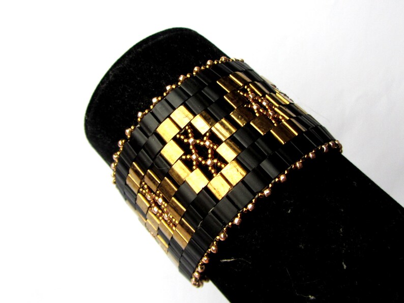 Op de afbeelding: Een zwarte en gouden kralen armband met een geometrisch patroon. De armband is gemaakt met kleine, vierkante kralen en is versierd met kleinere, ronde kralen.