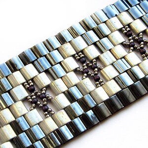 Op de afbeelding: Een zilveren en witte kralen armband met bruine kralen die een geometrisch patroon vormen.