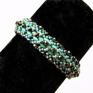 Pu&ograve; includere: Un braccialetto di perline turchesi e argento con un design intrecciato. Il braccialetto &egrave; esposto su un supporto in velluto nero.