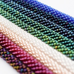 May include: A close-up of a variety of colorful seed beads, including silver, red, green, blue, and black. The beads are arranged in rows, creating a textured and iridescent pattern.