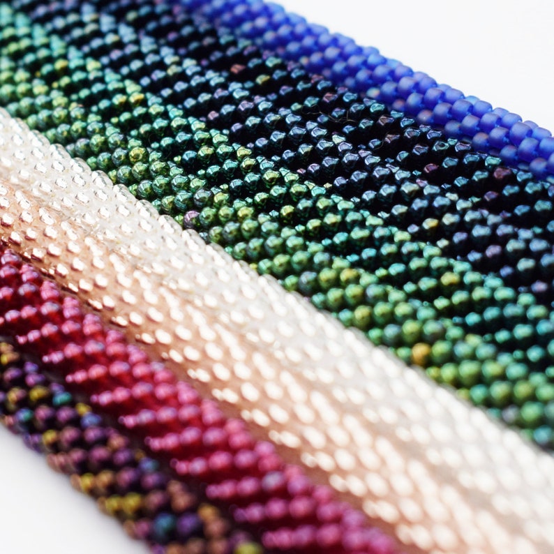 May include: A close-up of a variety of colorful seed beads, including silver, red, green, blue, and black. The beads are arranged in rows, creating a textured and iridescent pattern.