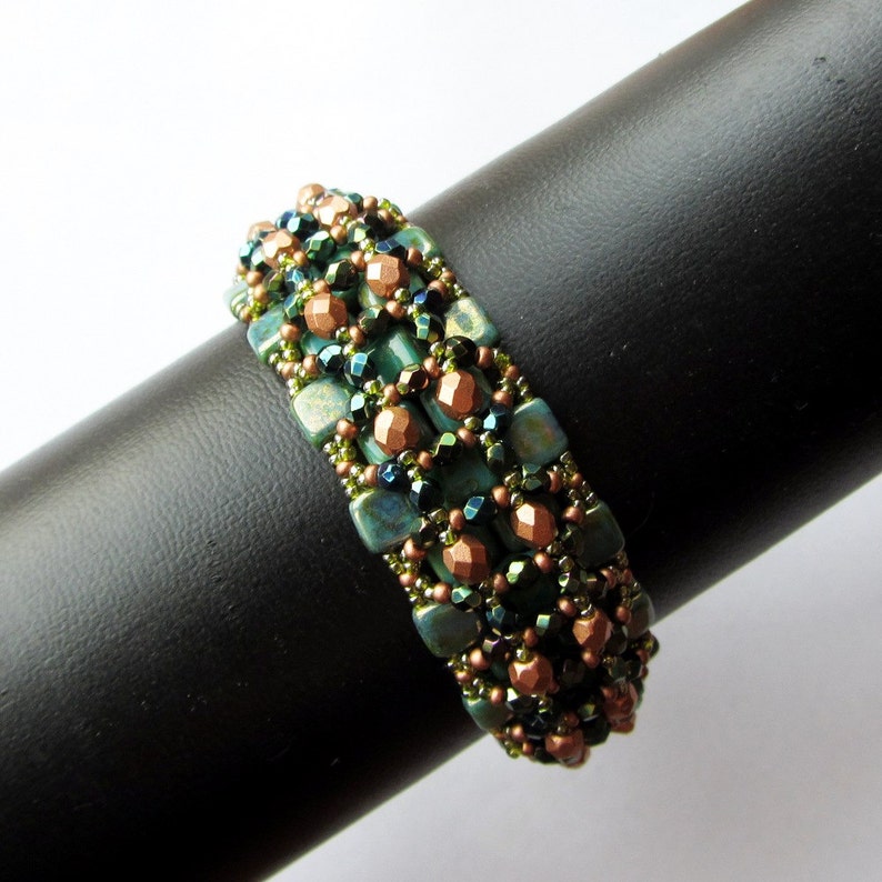 May include: A green and copper beaded bracelet with a geometric pattern. The bracelet is on a black display stand.