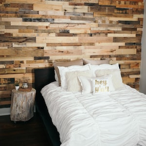 May include: A white bed with a white duvet and several pillows, including one with the text "hey y'all" in gold lettering. The bed is in front of a wall made of reclaimed wood planks.