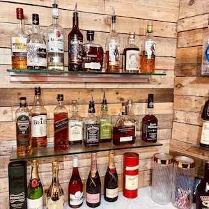 May include: A home bar with two shelves stocked with various bottles of spirits, including whisky, rum, and champagne. The shelves are made of wood and glass. There are also two glass jars with straws and a bottle of cream liqueur on the counter.