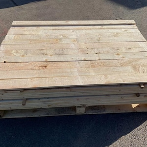 May include: A stack of unfinished wooden planks, possibly for a DIY project or construction. The planks are light brown and appear to be softwood. The stack is on a wooden pallet.
