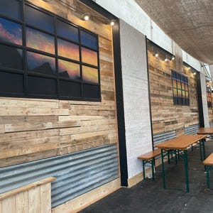 May include: A rustic outdoor dining area with wooden walls, a corrugated metal panel, and a white brick wall. There are two wooden tables with green metal legs and benches. The tables are set up in front of the walls.
