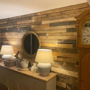 May include: A distressed wooden wall with a large, round mirror with a gold frame. Two lamps with white shades are on a white dresser with drawers. A grandfather clock with a wooden case and a white face is in the corner of the room.
