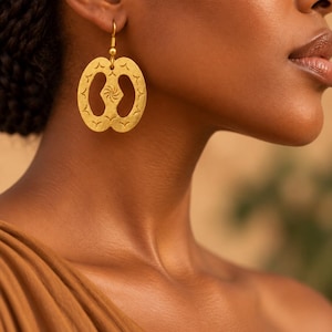 May include: A gold-toned dangle earring featuring a flat, oval-shaped pendant with a central cutout and a small spiral design. The surface of the pendant is textured with small, repeating V-shaped markings along the outer edge and a hook closure.
