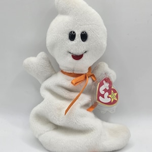 Può includere: Un giocattolo a forma di fantasma di peluche bianco con occhi a bottone neri e un sorriso cucito rosso. Ha un nastro arancione intorno al collo e un'etichetta attaccata. Il giocattolo è un oggetto da collezione.
