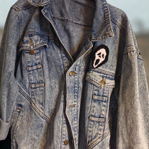 May include: Acid wash denim jacket with a black and white patch featuring a ghost face design. The jacket has two chest pockets, two side pockets, and gold-colored buttons. The sleeves are cuffed, and the jacket is hanging on a black hanger.