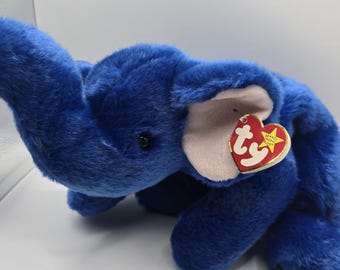 May include: A plush blue elephant toy with a trunk, ears, and a tag. The elephant is lying down on a white surface. The tag is red and yellow with the text "BEANIE BUDDY".
