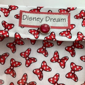 K&ouml;nnte beinhalten: Eine wei&szlig;e Stofftasche mit einem roten Polka-Dot-Schleifenmuster. Die Tasche hat ein rotes Etikett mit der Aufschrift "Disney Dream" und einen roten Knopfverschluss.