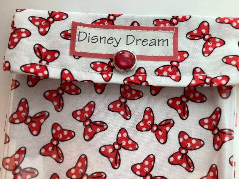 K&ouml;nnte beinhalten: Eine wei&szlig;e Stofftasche mit einem roten Polka-Dot-Schleifenmuster. Die Tasche hat ein rotes Etikett mit der Aufschrift "Disney Dream" und einen roten Knopfverschluss.