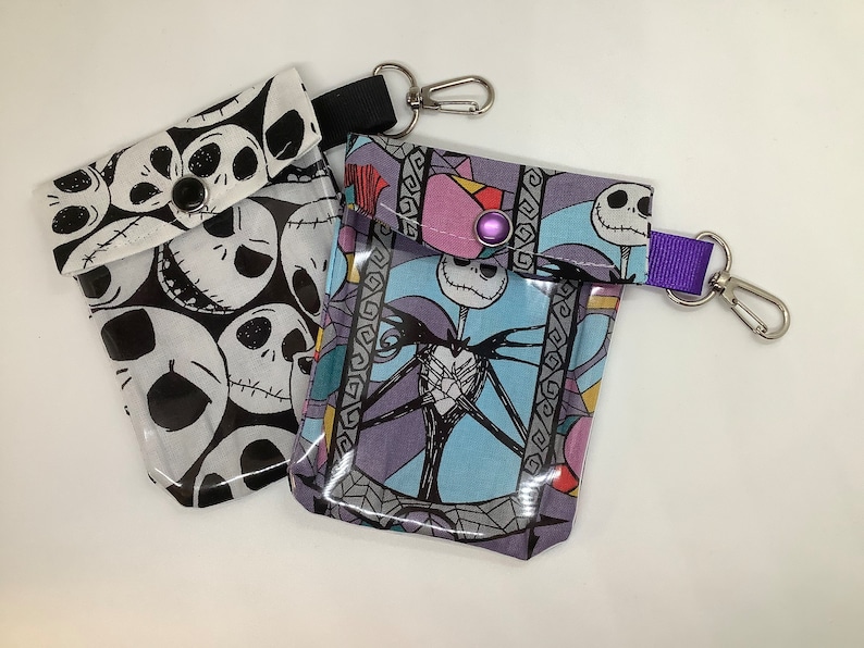 May include: Two small zippered pouches with a clear plastic window. One pouch is black and white with a repeating pattern of Jack Skellington's face. The other pouch is blue, purple, and white with a picture of Jack Skellington. Both pouches have a silver metal clip and a purple strap.