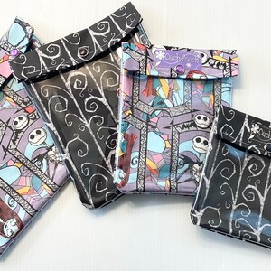 May include: Four clear plastic zipper pouches with different Nightmare Before Christmas fabric designs. The pouches are black with white swirls, black with colorful stained glass designs, and purple with colorful stained glass designs. The pouches have a clear plastic front and a black fabric back. The pouches have a small flap with a snap closure. The pouches have a small tag that says "Clutch Pouch".