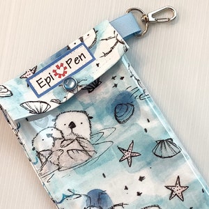 以下が含まれることがあります： 青いと白い布地のポーチで、透明なプラスチックの窓が付いています。アザラシのデザインと「Epi Pen」の文字が書かれています。ポーチには青いストラップと銀色のキーリングが付いています。