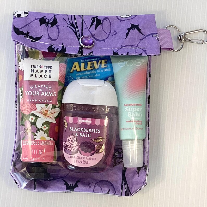 Puede incluir: Una bolsa de cremallera morada con un bolsillo frontal transparente que contiene varios art&iacute;culos de cuidado personal, como crema de manos, desinfectante de manos, b&aacute;lsamo labial y pastillas para el dolor. La bolsa tiene un patr&oacute;n de murci&eacute;lagos blanco y negro y un tirador de cremallera plateado.