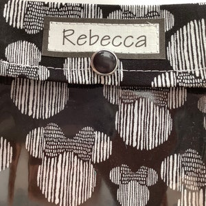 K&ouml;nnte beinhalten: Schwarze Stofftasche mit einem durchsichtigen Plastikfenster. Die Tasche hat ein wei&szlig;es Etikett mit dem Namen "Rebecca" darauf. Der Stoff hat ein sich wiederholendes Muster aus wei&szlig;en, stilisierten Kreisen auf schwarzem Hintergrund.