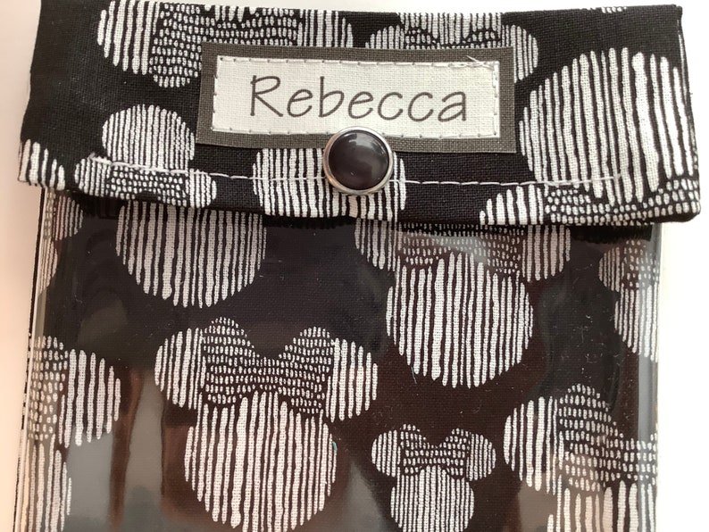 K&ouml;nnte beinhalten: Schwarze Stofftasche mit einem durchsichtigen Plastikfenster. Die Tasche hat ein wei&szlig;es Etikett mit dem Namen "Rebecca" darauf. Der Stoff hat ein sich wiederholendes Muster aus wei&szlig;en, stilisierten Kreisen auf schwarzem Hintergrund.