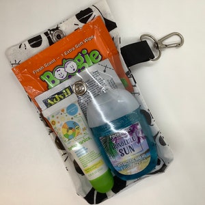 May include: A clear plastic pouch with a black and white patterned fabric exterior. The pouch contains a small bottle of hand sanitizer, a tube of sunscreen, a tube of lip balm, and a small packet of Boogie wipes. The pouch has a black strap with a silver clip.