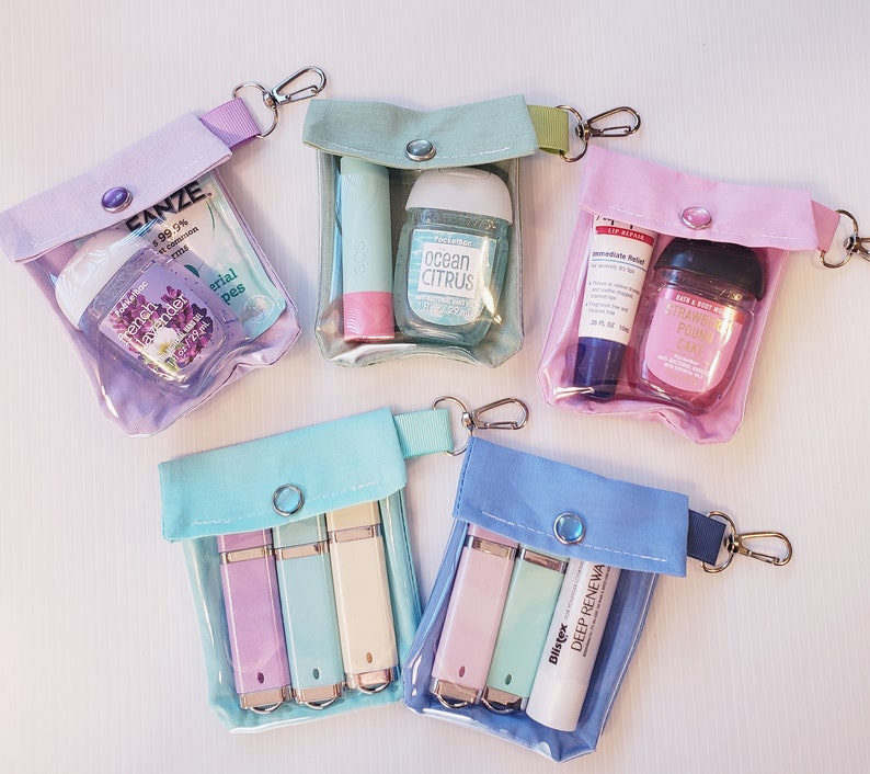 May include: Five clear plastic zippered pouches with different colored fabric exteriors. Each pouch contains various small items, including hand sanitizer, lip balm, and small tubes. The pouches have a zipper closure and a metal ring for attaching to a keychain.