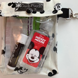 K&ouml;nnte beinhalten: Eine wei&szlig;e Stofftasche mit einem durchsichtigen Plastikfenster, bedruckt mit einem schwarz-wei&szlig;en Mickey Mouse-Motiv. Die Tasche hat einen schwarzen Knopfverschluss und einen schwarzen Riemen mit einem silbernen Karabinerhaken. In der Tasche befinden sich ein R&ouml;hrchen mit ChapStick Lippenbalsam, eine kleine Flasche Mickey Mouse H&auml;ndedesinfektionsmittel und ein kleines P&auml;ckchen mit Salzl&ouml;sung f&uuml;r die Nase.