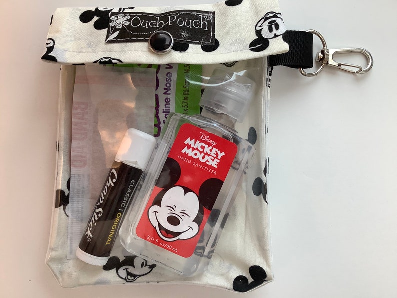 K&ouml;nnte beinhalten: Eine wei&szlig;e Stofftasche mit einem durchsichtigen Plastikfenster, bedruckt mit einem schwarz-wei&szlig;en Mickey Mouse-Motiv. Die Tasche hat einen schwarzen Knopfverschluss und einen schwarzen Riemen mit einem silbernen Karabinerhaken. In der Tasche befinden sich ein R&ouml;hrchen mit ChapStick Lippenbalsam, eine kleine Flasche Mickey Mouse H&auml;ndedesinfektionsmittel und ein kleines P&auml;ckchen mit Salzl&ouml;sung f&uuml;r die Nase.