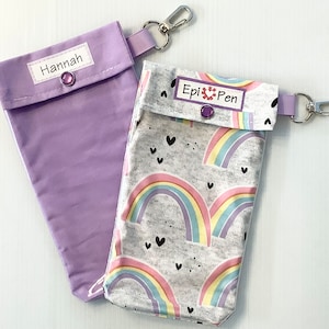 May include: Two purple fabric pouches with zipper closures and a snap button. One pouch has the name "Hannah" printed on it. The other pouch has a rainbow and heart pattern and the text "Epi Pen" printed on it.