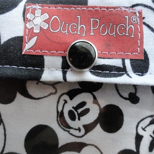 May include: Close-up of a fabric pouch with a black and white Mickey Mouse pattern. The pouch has a red label with the text "Ouch Pouch" and a black snap closure. The background is a white fabric with black Mickey Mouse faces.