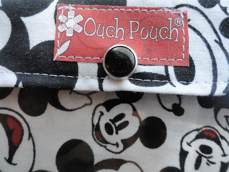 May include: Close-up of a fabric pouch with a black and white Mickey Mouse pattern. The pouch has a red label with the text "Ouch Pouch" and a black snap closure. The background is a white fabric with black Mickey Mouse faces.