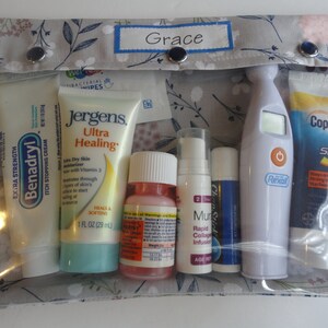 May include: A clear zippered pouch with a gray floral pattern and the word "Grace" on the front. The pouch contains various items, including tubes of lotion, a bottle of liquid, a thermometer, and a lip balm.
