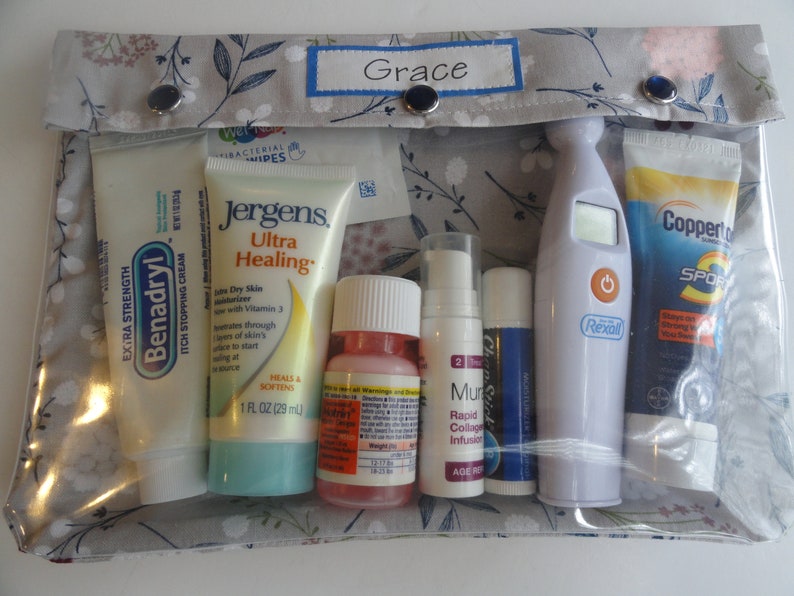 May include: A clear zippered pouch with a gray floral pattern and the word "Grace" on the front. The pouch contains various items, including tubes of lotion, a bottle of liquid, a thermometer, and a lip balm.
