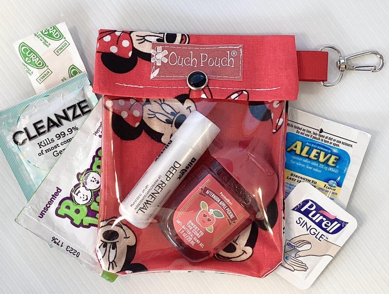 May include: Red zippered pouch with a clear plastic window, featuring a Mickey Mouse design. The pouch contains various items, including hand sanitizer, pain relief tablets, and a small tube of hand cream. The pouch has a red strap with a silver carabiner clip.