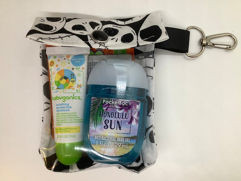 May include: A clear plastic zippered pouch with a black and white skull print. The pouch contains a small green and white tube of babyganics soothing protective ointment and a blue bottle of PocketBac Honolulu Sun anti-bacterial hand gel. The pouch has a black strap with a silver clip.