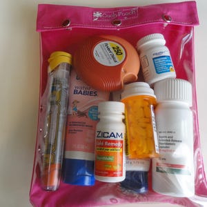 May include: A clear, pink, zippered pouch labeled "Cach Pouch" containing various medications. Items include an inhaler, a pre-filled epinephrine auto-injector, bottles of pills, and a tube of sunscreen. The pouch is closed with snaps.