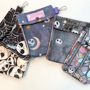 Puede incluir: Cuatro bolsas de tela diferentes con una ventana de pl&aacute;stico transparente. Cada bolsa presenta un dise&ntilde;o diferente con un personaje de Jack Skellington en blanco y negro de la pel&iacute;cula Pesadilla antes de Navidad. Las bolsas tienen una correa morada y un cierre de metal. El texto en las bolsas dice "Ouch Pouch", "Disney Dream" y "The Pumpkin King".